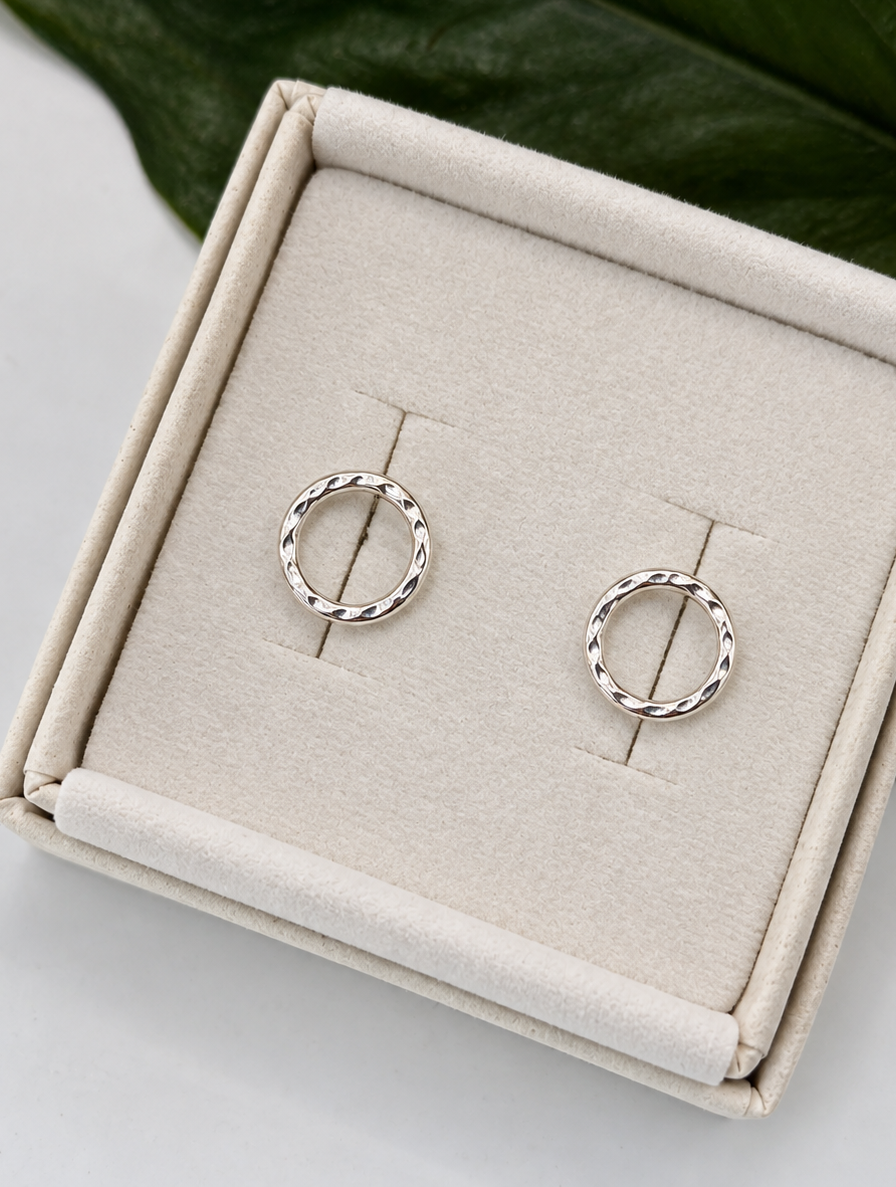 Pair of handmade recycled 925 sterling silver circle stud earrings with a delicate dimple texture, displayed on a black jewellery stand against a soft neutral background with a green leaf.